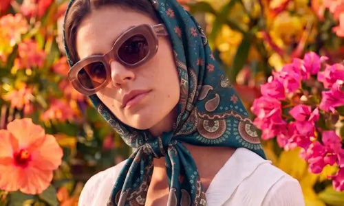 Imagem de mulher com o lenço Oriental Astrid azul na cabeça usando óculos em ambiente de flores.