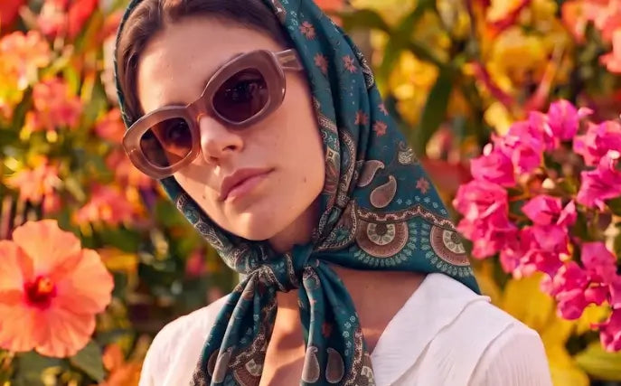 Imagem de mulher com o lenço Oriental Astrid azul na cabeça usando óculos em ambiente de flores.