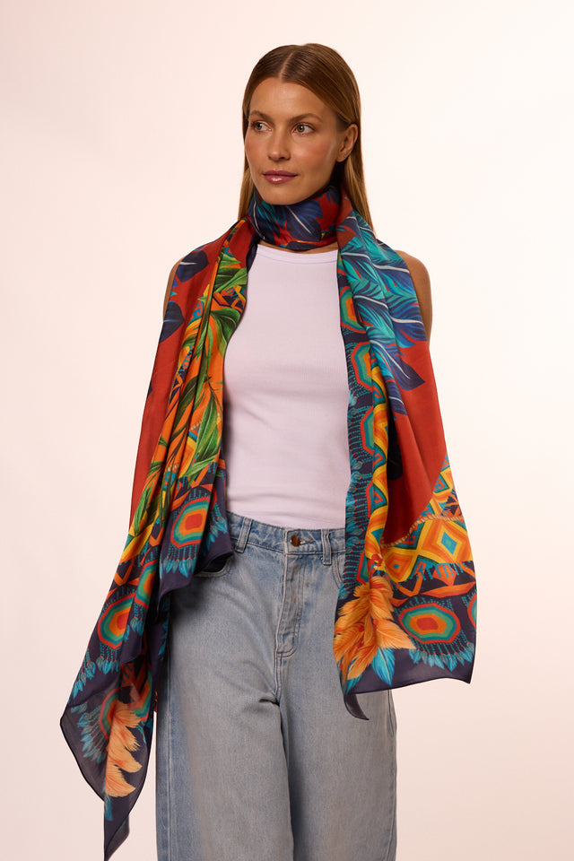 Mulher com cabelo castanho claro, usando camiseta branca e jeans, com lenço no pescoço colorido com padrão vibrante, posando contra fundo bege.