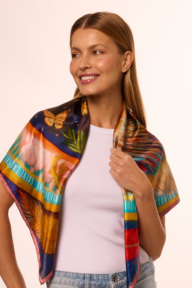 Modelo usando um lenço no pescoço colorido com borboletas, flores e penas. Sorriso confiante e look casual, simbolizando elegância e criatividade na moda.