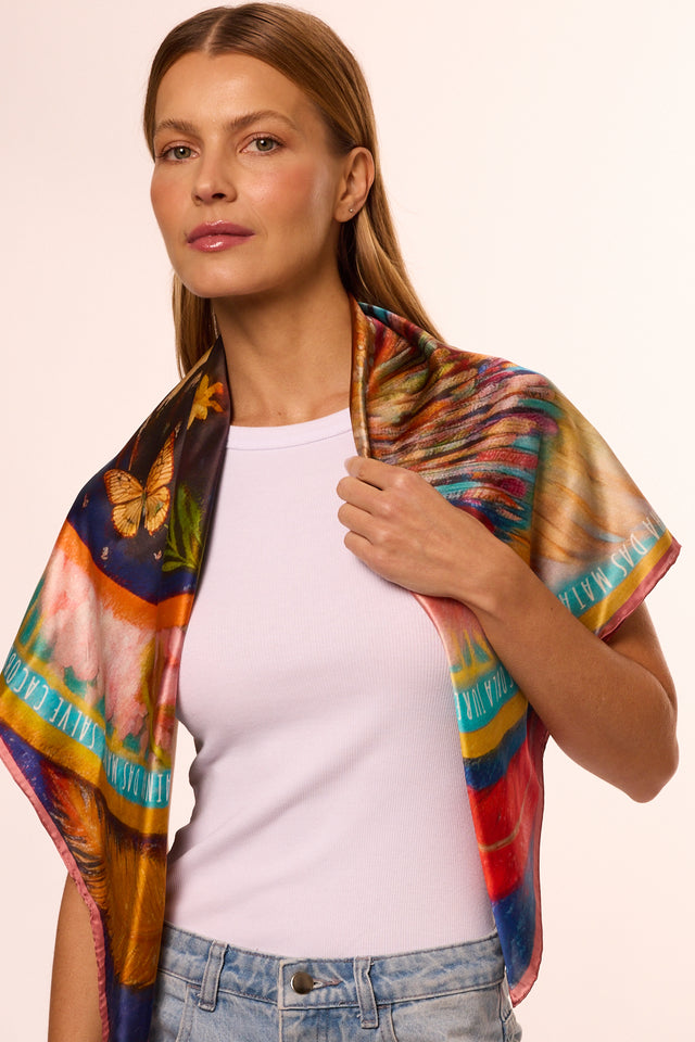 Modelo usando o lenço para pescoço Brilho de Jurema, da Scarf Me, com detalhes de borboletas e penas. Ela também veste regata branca e jeans azul, pose confiante e elegante.