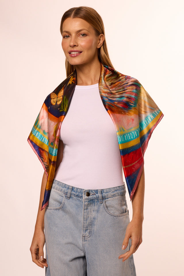 Mulher com cabelo castanho claro, usando blusa branca e calça jeans, com lenço no pescoço colorido com estampa de borboleta e outros desenhos artísticos, sorrindo em fundo claro.