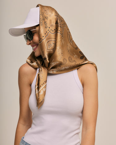 Mulher com lenço no pescoço marrom estampado, boné branco e óculos escuros sorrindo, vestindo camiseta branca, em fundo neutro, estilo casual e elegante.