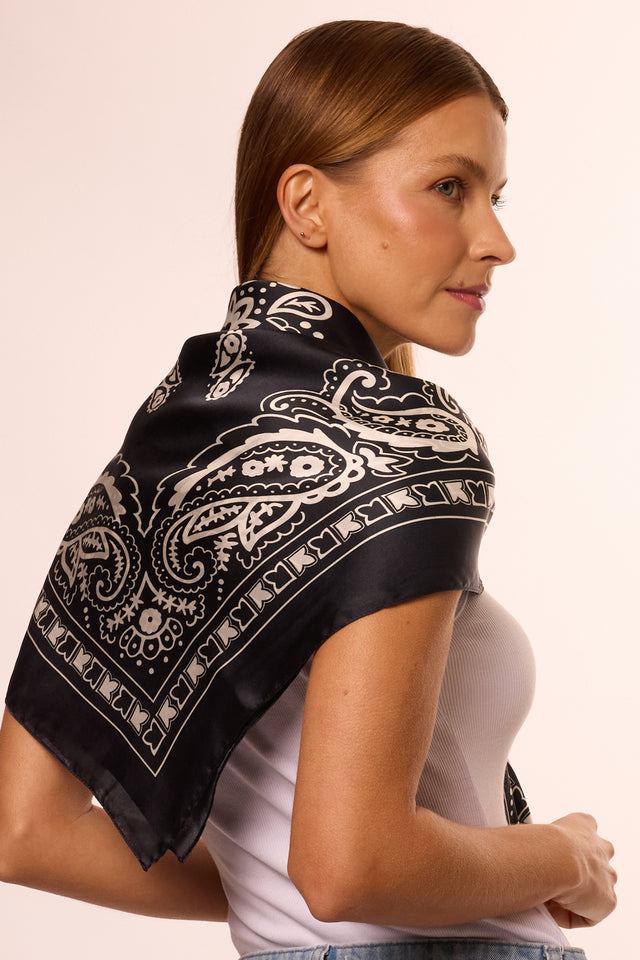 Modelo com cabelo castanho, usando blusa branca e lenço preto com estampa branca ao redor do pescoço, ela está de perfil em fundo branco.