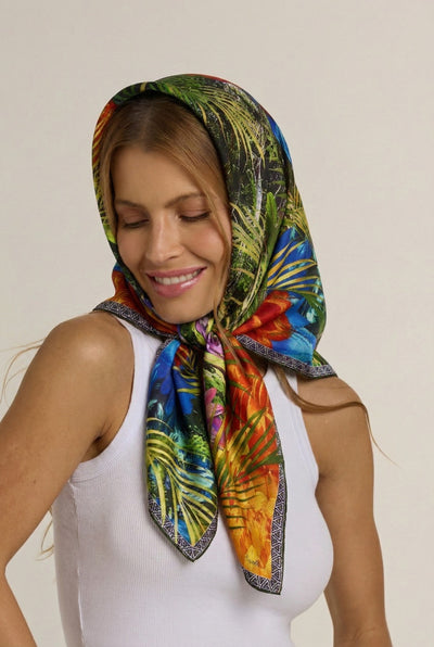 Mulher sorrindo com lenço no pescoço colorido sobre a cabeça, vestindo blusa branca, contra fundo neutro.