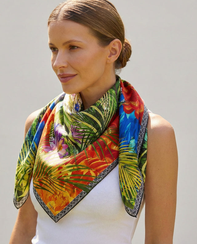 Mulher elegante usando lenço no pescoço com estampa de folhagem e flores coloridas, com cabelo preso e expressão suave, contra fundo claro.