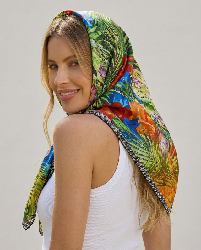 Mulher sorridente com cabelo castanho, vestindo camiseta branca e usando um lenço no pescoço colorido com estampa de folhas e flores.