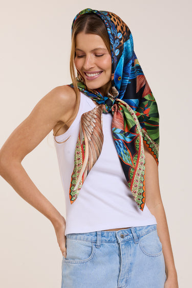 Mulher sorridente usando lenço colorido no cabelo e roupa casual, com destaque para o lenço de seda vibrante e estilo descontraído.