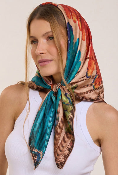 Mulher com cabelo castanho claro usando lenço de seda colorido com padrão de penas, regata branca e feições delicadas, posando contra fundo neutro.