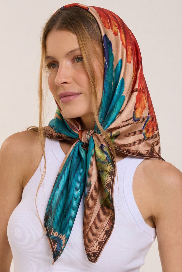Mulher com cabelo castanho claro usando lenço de seda colorido com padrão de penas, regata branca e feições delicadas, posando contra fundo neutro.