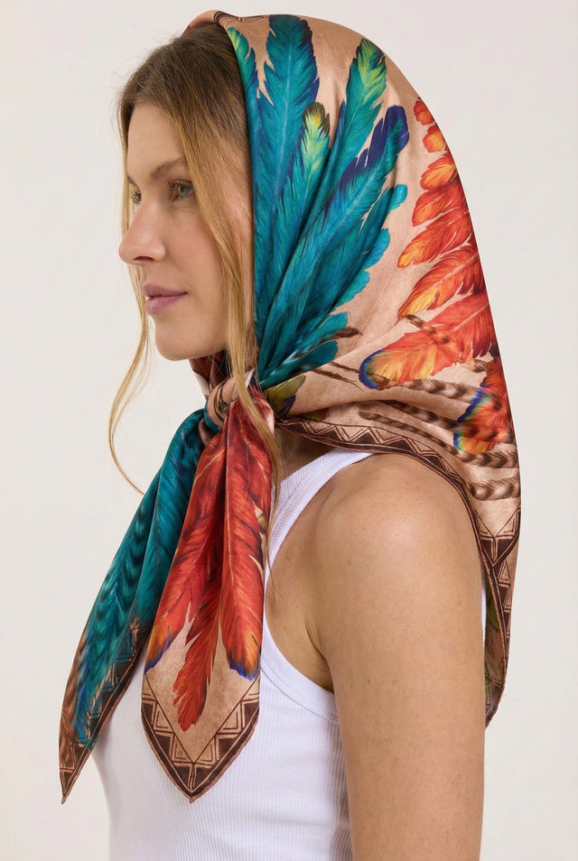 Mulher com cabelos castanhos claros usando lenço de seda com estampa de penas coloridas em tons de azul, vermelho e laranja, de perfil, contra fundo branco.