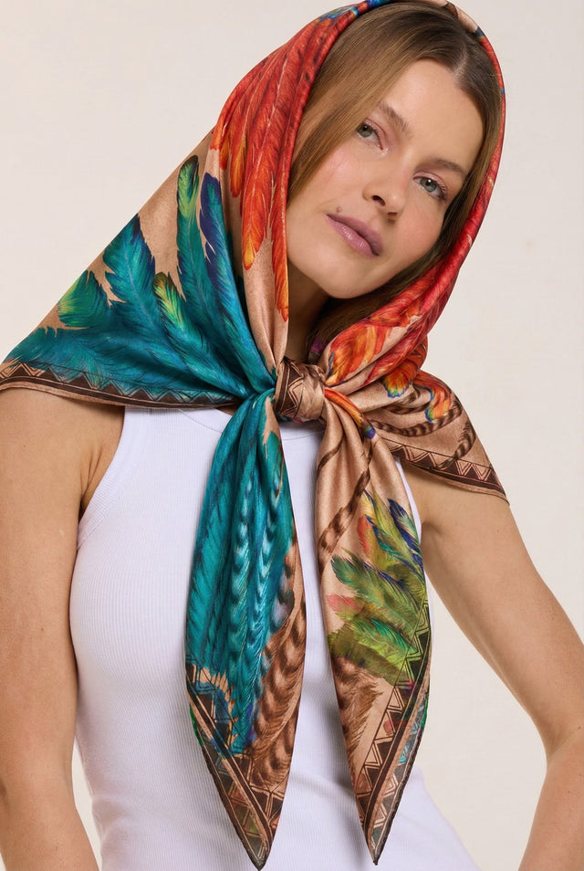 Mulher com lenço colorido estampado sobre a cabeça amarrado no pescoço, usando blusa branca, com cabelo castanho claro e expressão serena, contra fundo branco.