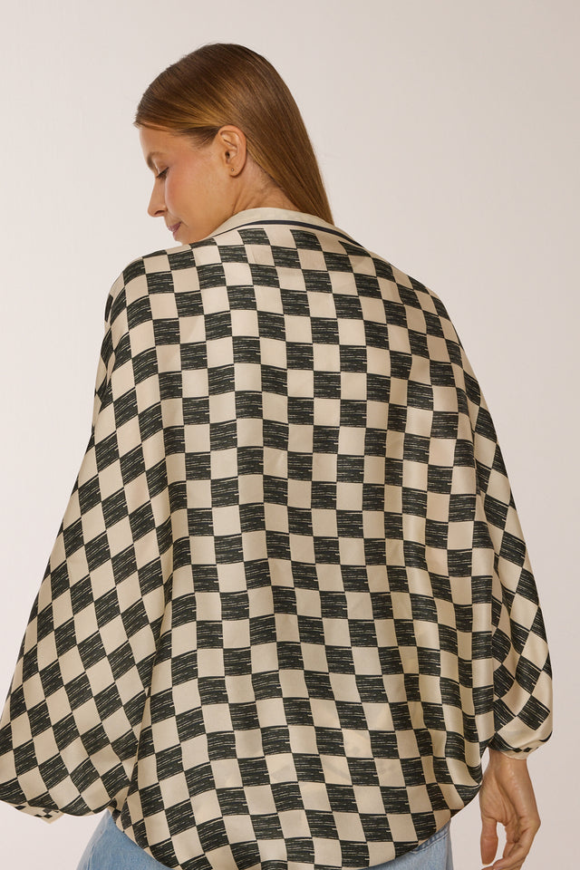 Imagem de modelo usando o lenço Race nos ombros, estampa quadriculada preta e branca, voltada de costas, em fundo neutro.