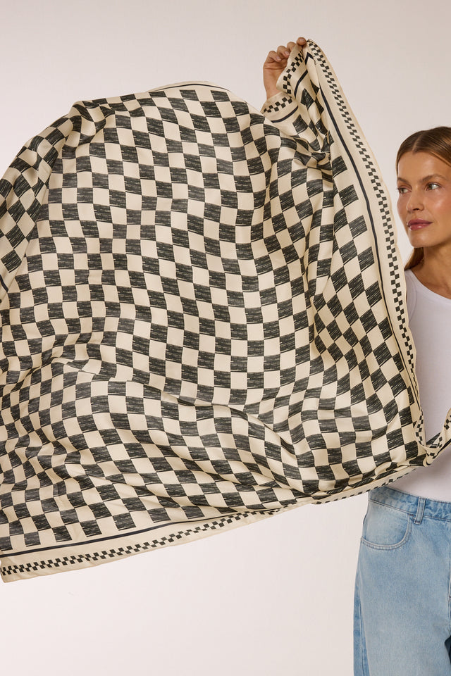 Mulher o lenço para pescoço Race, com estampa de xadrez em preto e branco, usando uma camiseta branca e jeans, em fundo claro.