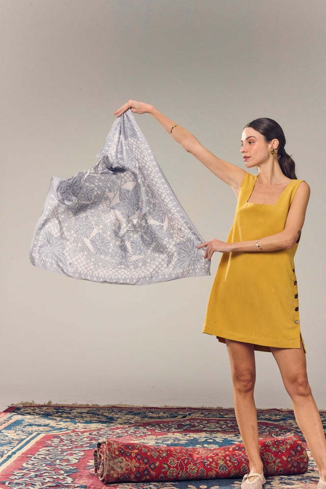 Mulher com vestido amarelo segurando lenço para pescoço cinza com padrões florais e geométricos, em um cenário simples com tapete vermelho e fundo neutro.