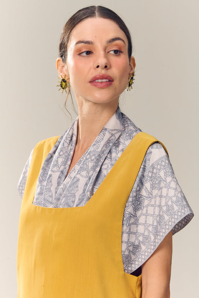 Mulher elegante com vestido amarelo, lenço ritos do mar vestido como blusa e jóias chamativas, olhando para o lado em fundo neutro, estilo fashion e contemporâneo.