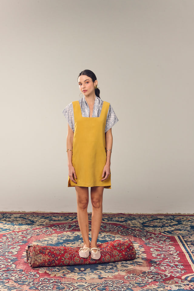 Mulher com cabelo preto, vestindo o lenço ritos do mar por baixo de vestido amarelo, em pé sobre um tapete vermelho e com fundo neutro.