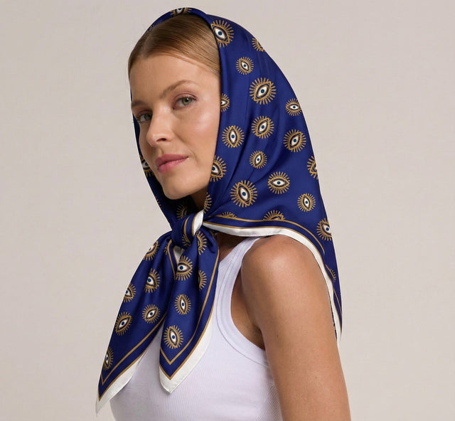 Mulher com lenço azul estampado com olho grego, vestindo regata branca, olhando de lado com expressão serena, fundo neutro.