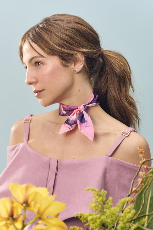 Modelo com lenço no pescoço Vestígios, em cetim de seda. Seu cabelo castanho est preso e ela veste camisa de alça na cor lilás.