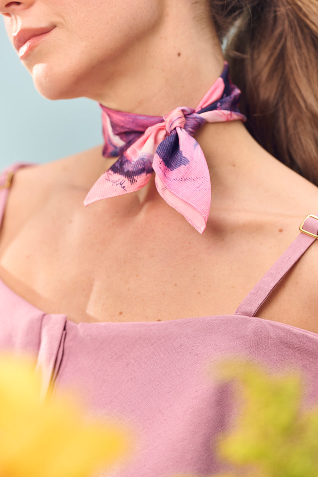 Lenço Vestígios amarrado em pescoço de modelo. A mulher posa em fundo azul e veste blusa de alça na cor lilás.